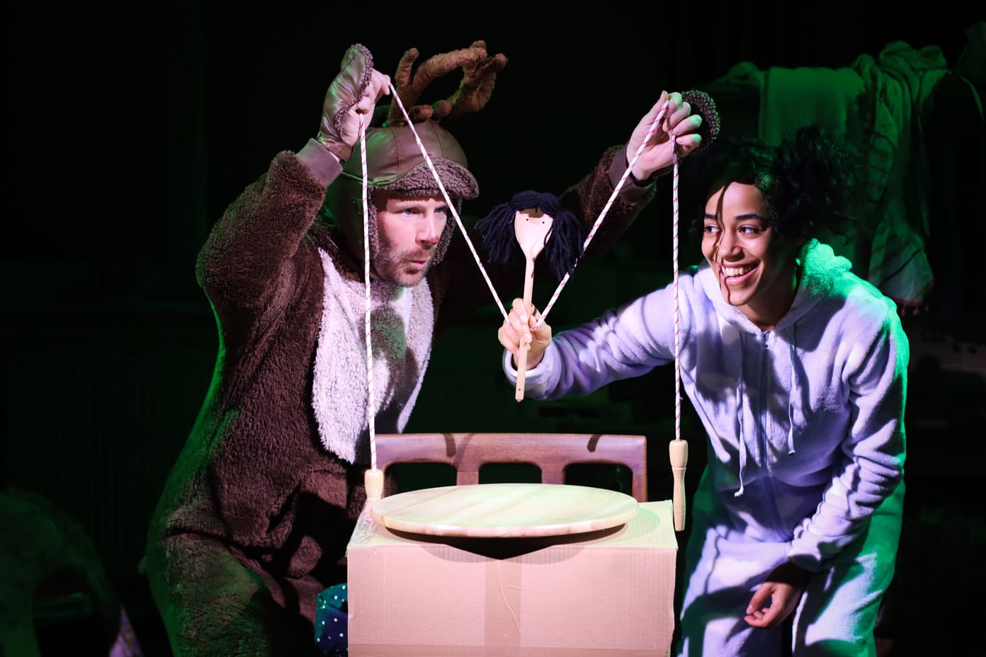 Two performers on stage in The Night Before Christmas operate a puppet made from a wooden spoon and a skipping rope. One is dressed as a reindeer, the other wears a blue onesie.