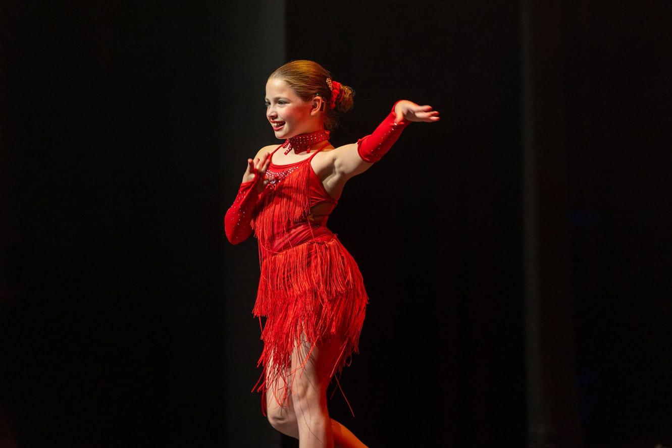 A smiling young performer on stage dances in a red tassled dress