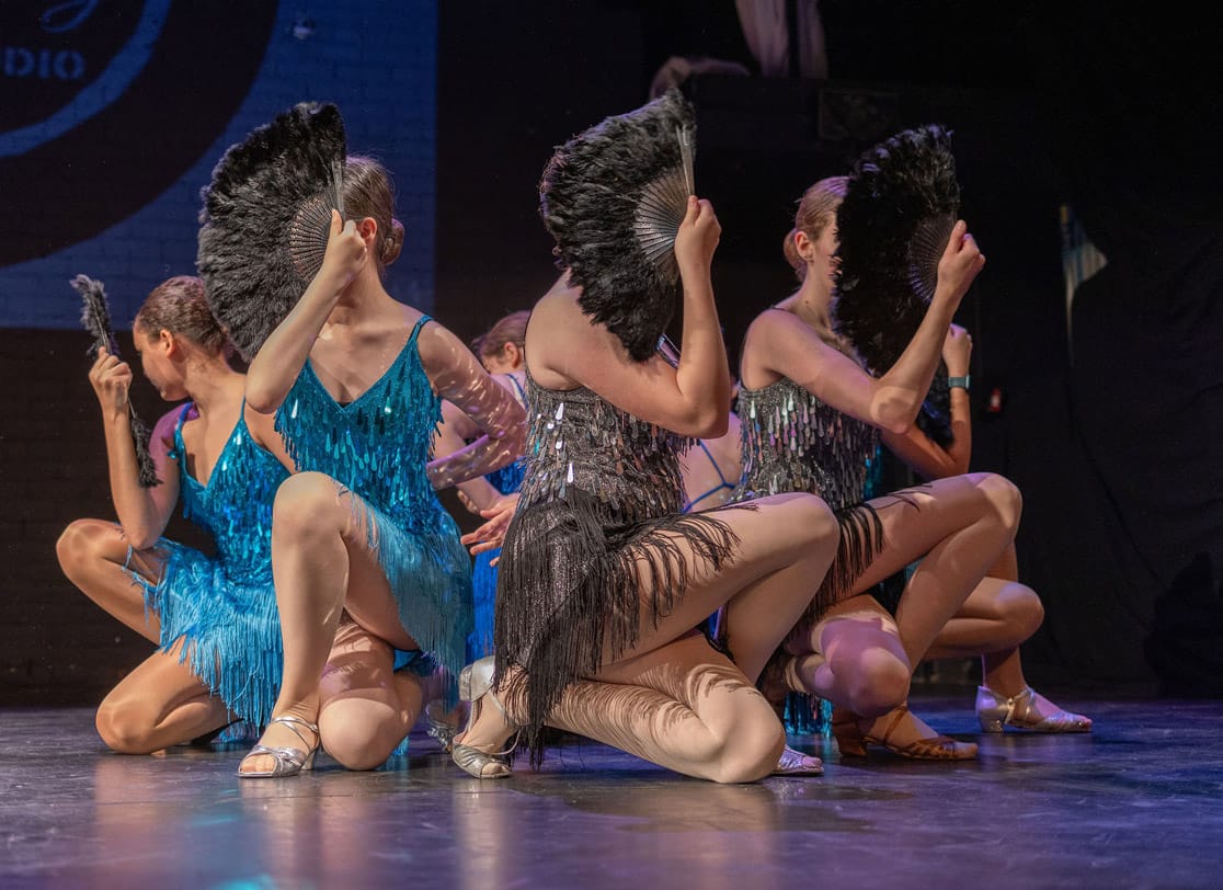 A group of young performers wearing blue and sliver tassled dresses hide their faces with grey feathered fans