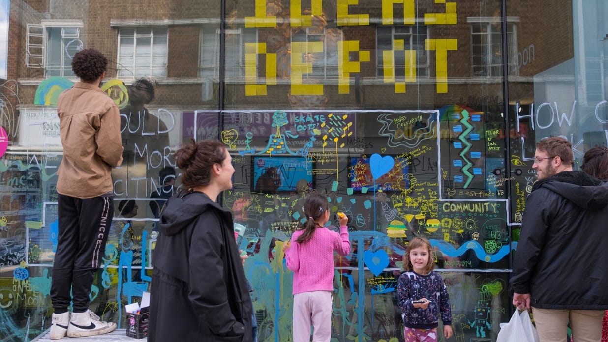 People of all ages draw and paint on artsdepot's windows. Ideas Depot is written with yellow post it notes at the top of the window.