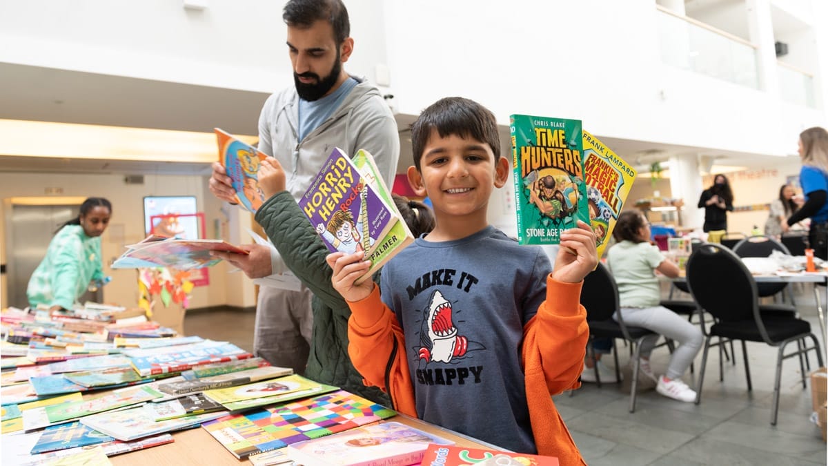 A child standing beside a table of books holding 4 books up and smiling