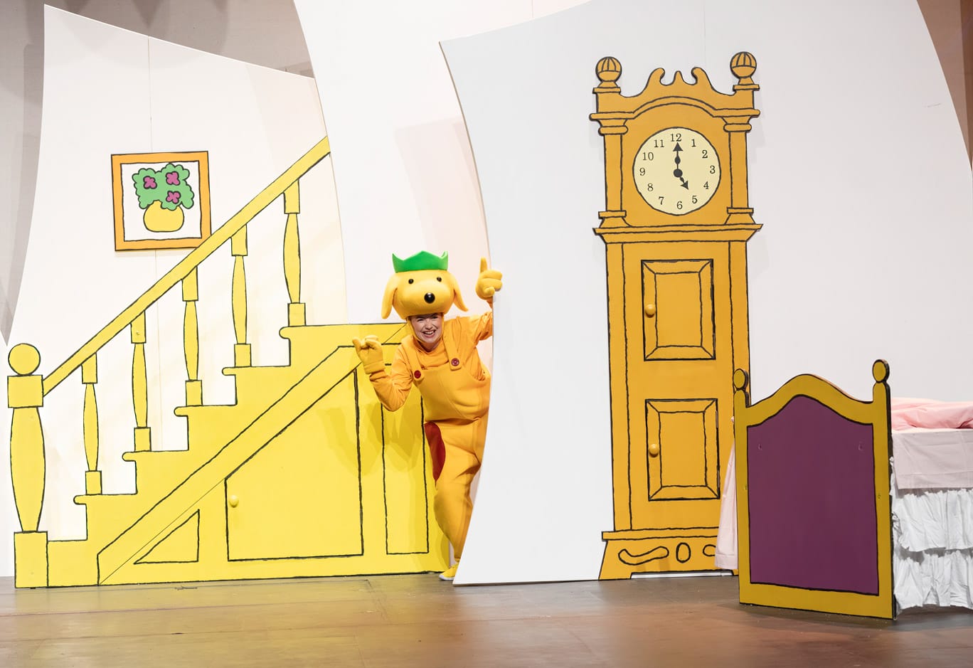 A performer on stage in Spot's Birthday Party. They are dressed as Spot the Dog with yellow dungarees and a yellow dog head on top of their head. They are waving from behind part of the set, which is illustrated to look like the inside of a house.