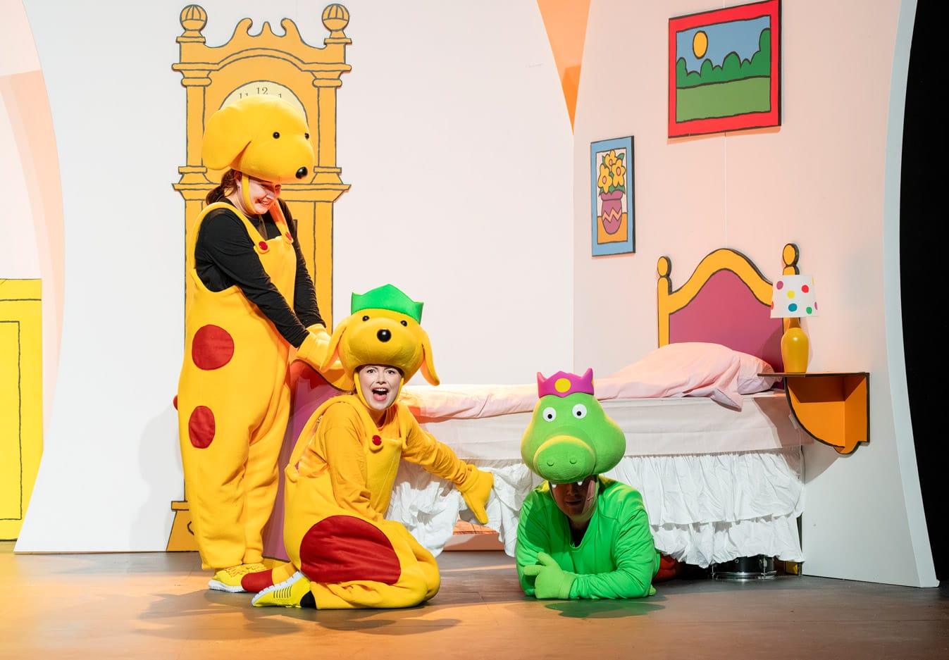 Performers on stage in Spot's Birthday Party, two wear dog costumes and the other, wearing a crocodile costume, is lying underneath a bed.