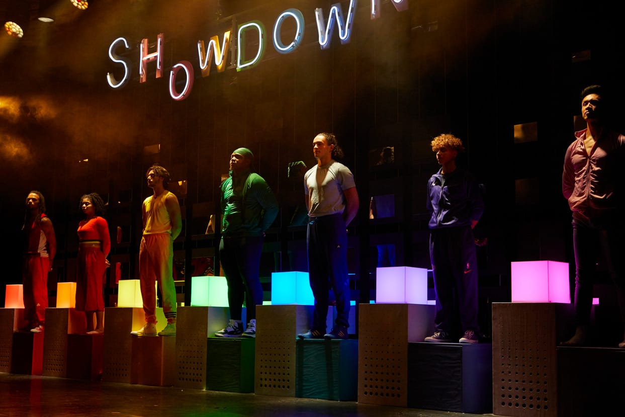 7 performers on stage in Showdown. They are standing in a line on blocks, with square lights behind them in the colours of the rainbow.
