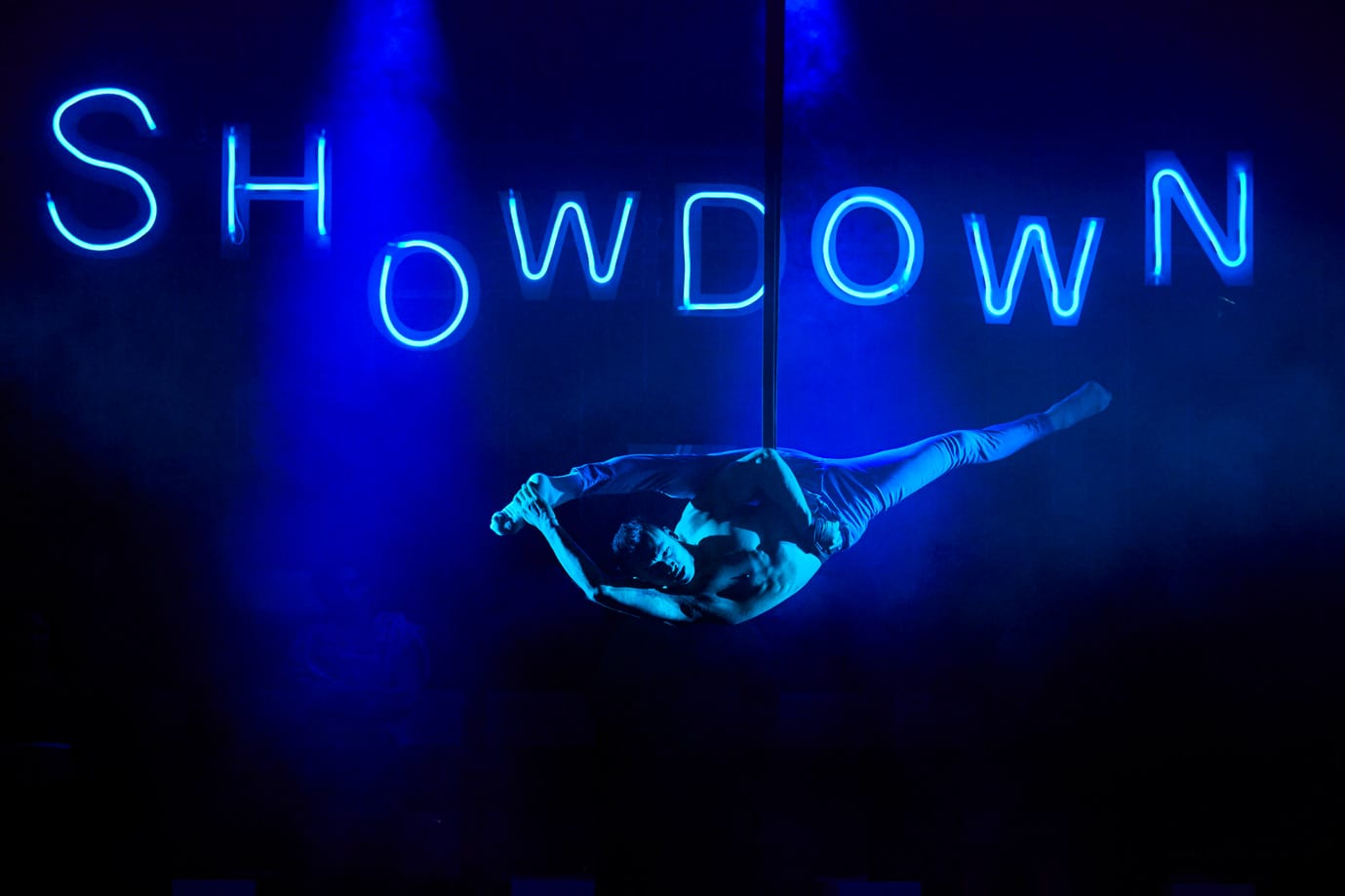 An aerial performer in Showdown. He is hanging from a rope with one leg stretched above his head and lit by blue light. Behind him, Showdown is spelled out in blue neon letters.