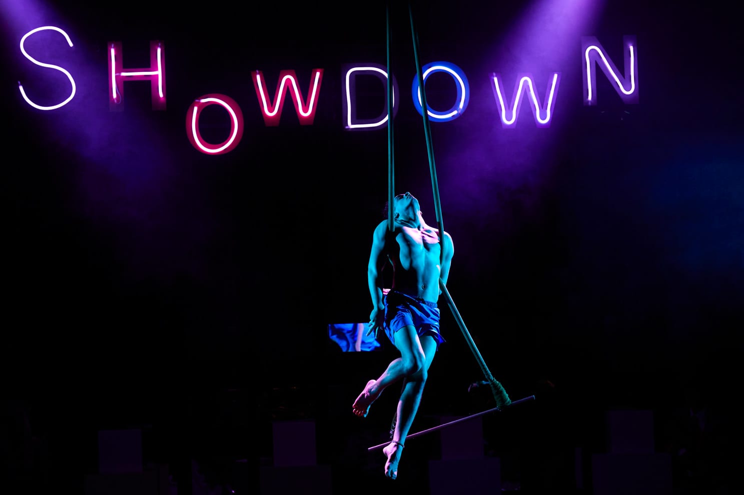 An aerial performer in Showdown hangs from a swing with their head thrown back. Neon lights behind them spell out Showdown.