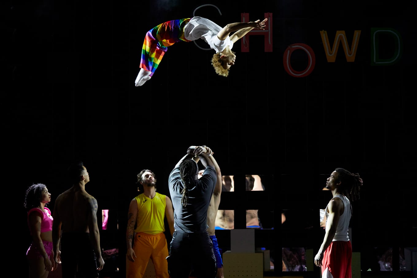 An acrobat flips through the air while another waits to catch him, four other performers watch on.
