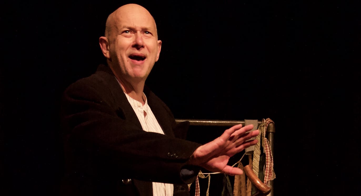 Bald man in a black suit, gesturing with his hand on stage in front of a black backdrop.