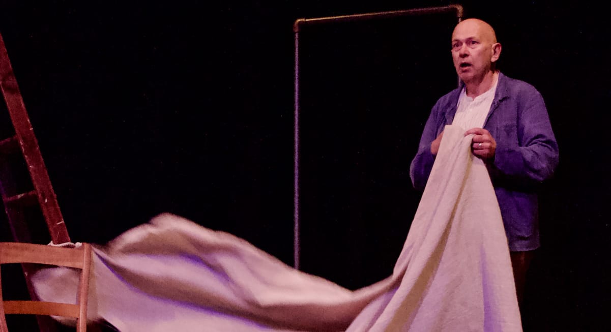 On a dimly lit theatre stage, a bald middle-aged actor, wearing a white shirt and a blue jacket, holds a tan coloured cloth.