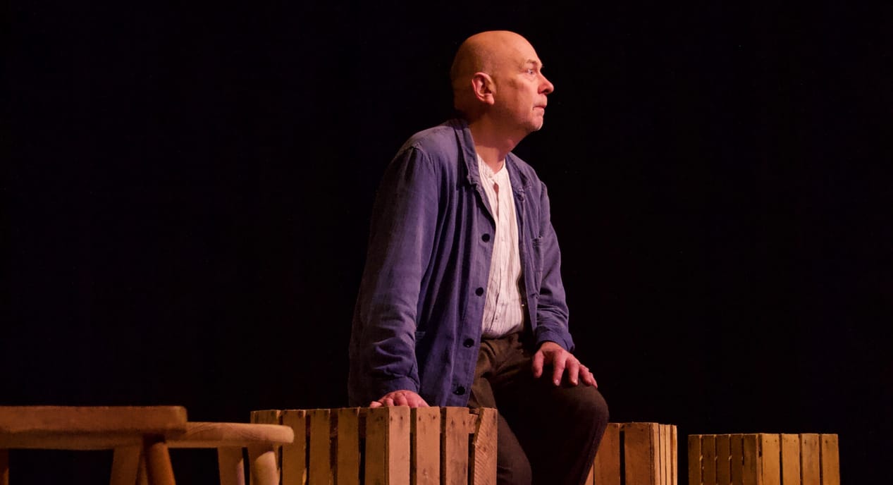 Man in jacket sits on wooden crates, looking off to the side against a black background.