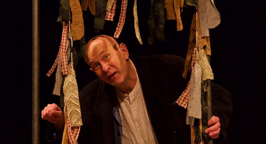 Actor in a play, peering through fabric strips. He wears a striped shirt and a dark jacket, against a black backdrop.