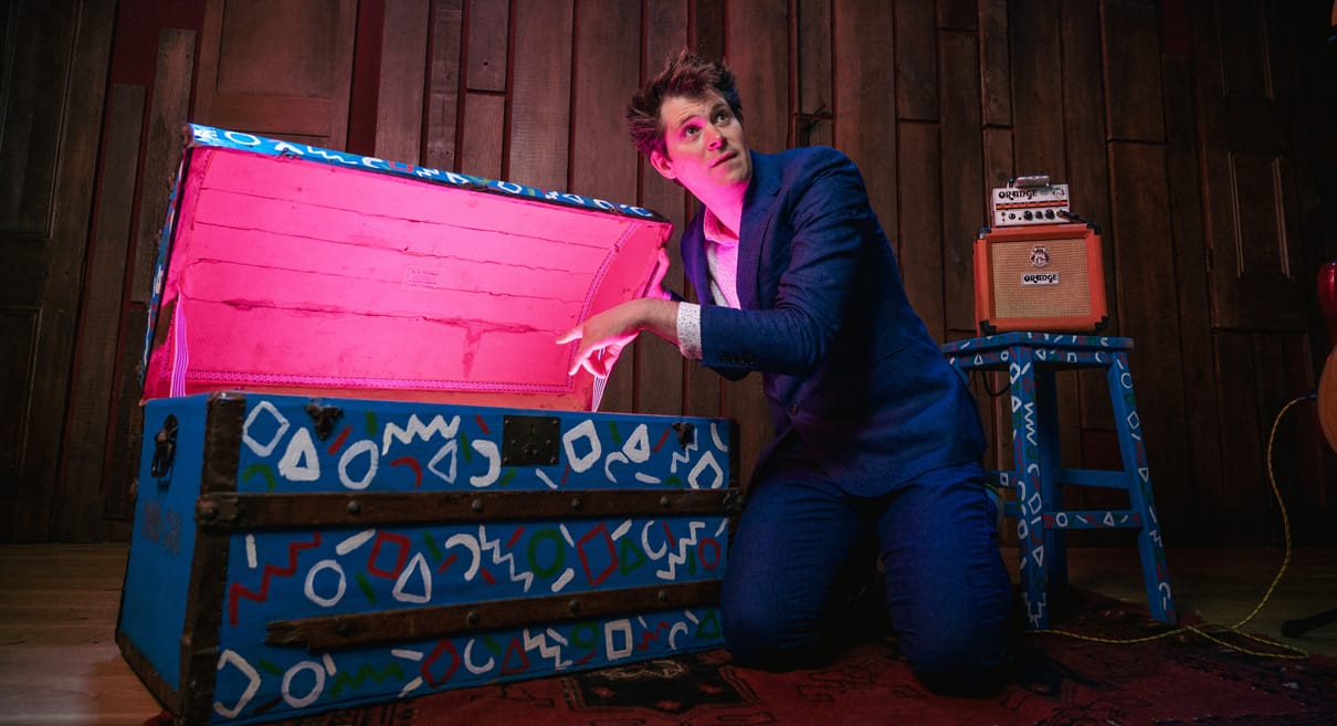 David Gibb at his Family Jukebox show, kneeling by an open trunk. The trunk interior glows pink.