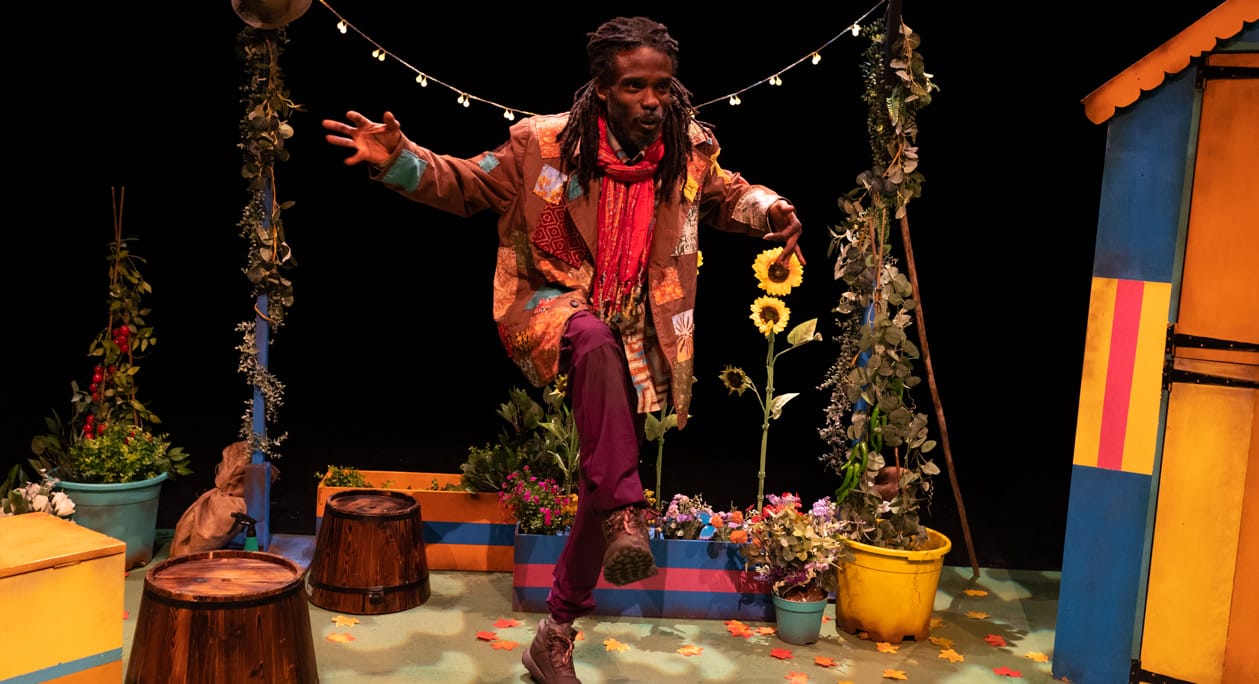 A black actor wearing a colourful, patterned jacket and a red scarf. The stage is set with faux flowers, sunflowers, painted boxes and wooden barrels, creating a vibrant garden setting, against a black background. A strand of lights is hanging above the actor.