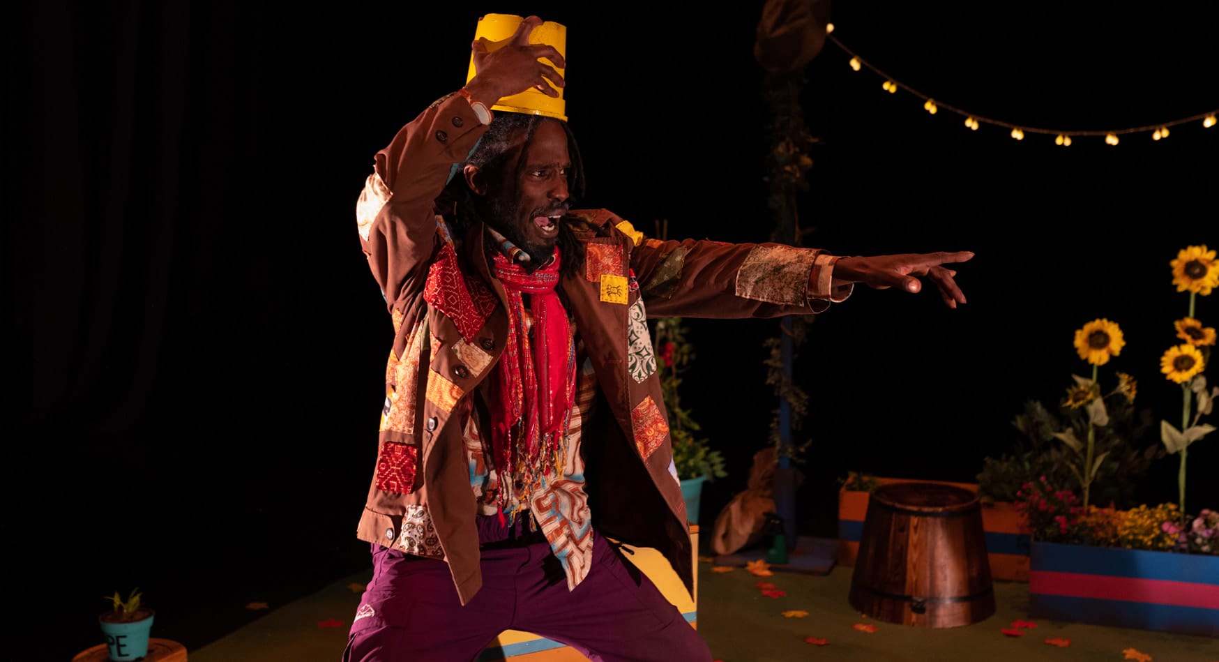 A Black actor with dreadlocks, wearing a tattered brown patchwork jacket, red scarf, and purple pants, points his right arm out dramatically into the audience. He holds a yellow bucket on his head. The stage backdrop is black, with light string, sunflowers and greenery on stage.