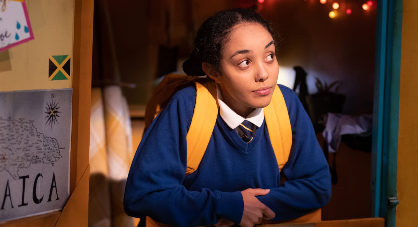 A young girl is resting her hands on a counter and looking thoughtfully off-camera. A wall to her left has a map of Jamaica and a Jamaican flag. A string of lights is visible in the background.