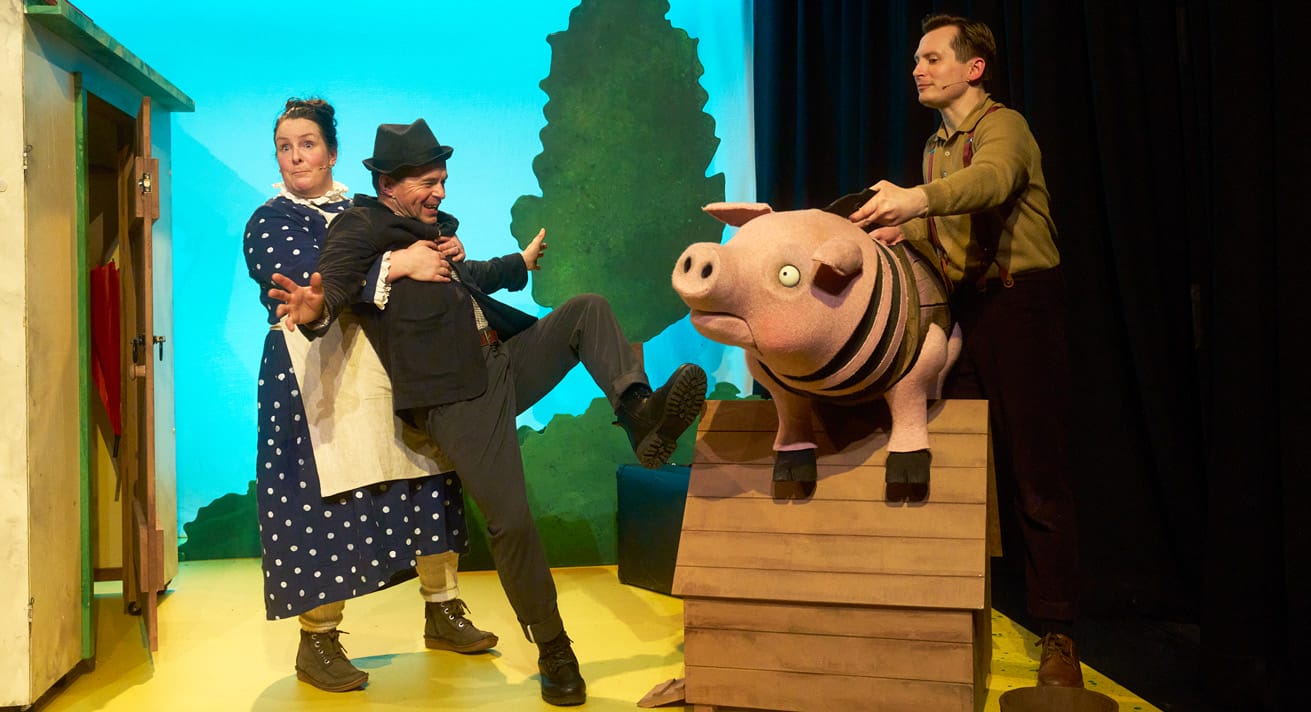 A production shot from A Squash and A Squeeze. A woman wearing a blue polka dot dress and white apron stands with her arms outstretched, looking on at a man in a black suit who is flailing slightly, his arms above and his legs up. An actor holding a pig puppet stands to the right.