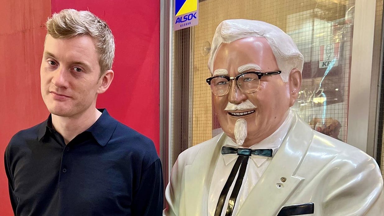 Comedian James Acaster standing next to a life size model of Colonel Sanders