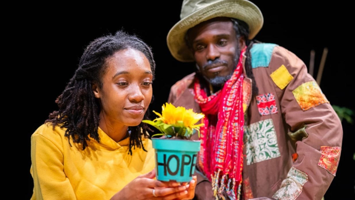 A close-up of two Black people looking at a small teal flowerpot with a yellow flower, which has the word 
