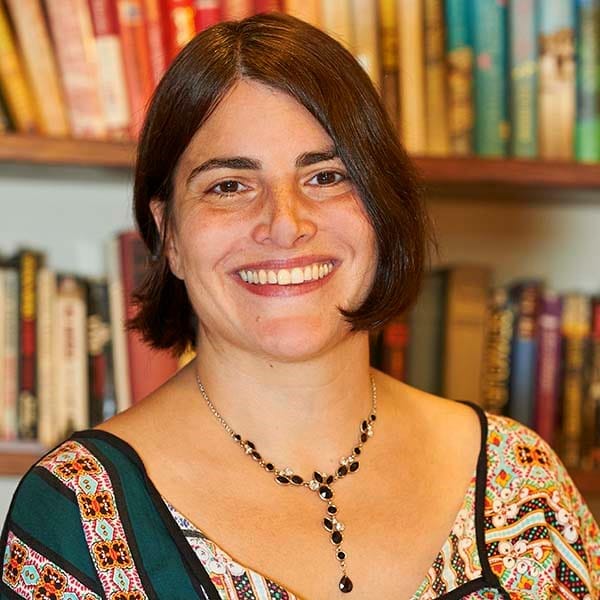 A photo of Creative Circle tutor Nichola Charalambou, a smiling woman with chin-length brown hair, a patterned top and a necklace. She is pictured in front of bookshelves.