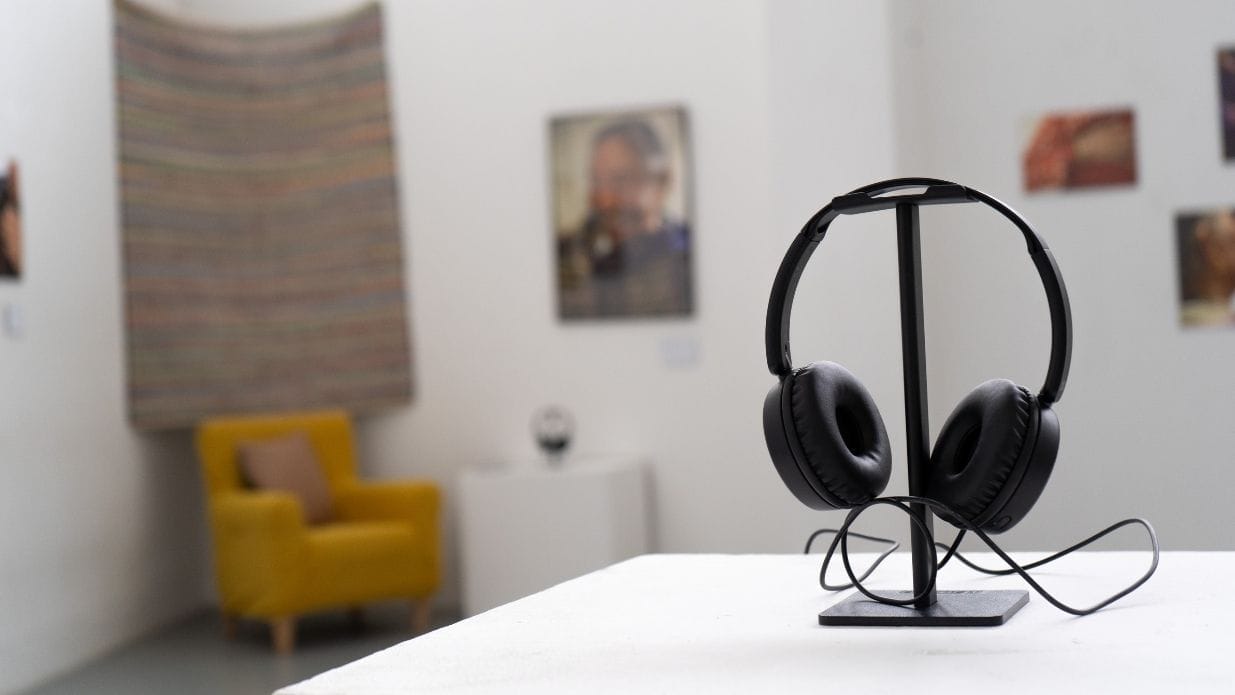 The interior of artsdepot's Apthorp Gallery. In the foreground a pair of headphones on a stand. In the background can be seen a yellow armchair and a rug, and large scale photographs displayed on the wall.