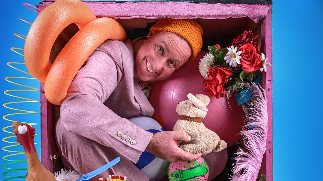A man in a pastel pink suit poses inside a box packed with various toys, an exercise ball and fake flowers.