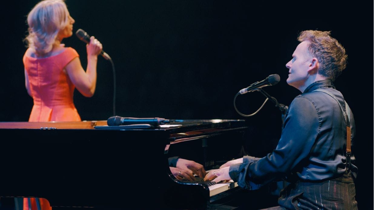 Joe Stilgoe sings at a black grand piano in the foreground. Behind him, Liza Pulman is singing wearing an orange dress.