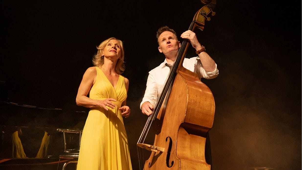 Liza Pulman and Joe Stilgoe are standing on stage and photographed slightly from below. Joe is holding a double bass.