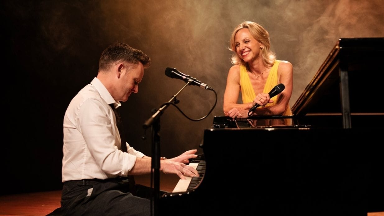 Liza Pulman smiles at Joe Stilgoe as he plays the piano.