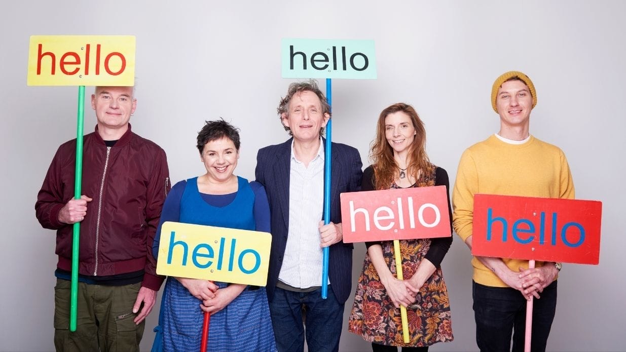 5 Members of Mrs H and the Sing-along band stand next to each other, each holding a sign that says 'hello'
