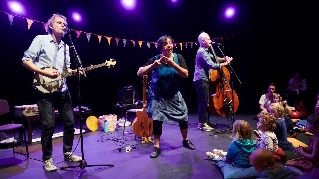 Mrs H and the Sing-along band on stage. Mrs H stands in the middle making a sign with her fingers. Next to her are a banjo player and a double bassist. A group of children are watching them.