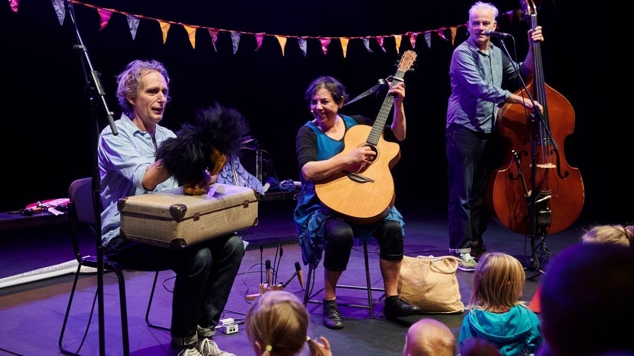 Mrs H and the Sing-along band on stage. Mrs H sits in the middle playing a guitar, either side of her are a standing double bassist and a seated member of the band holding a furry puppet.