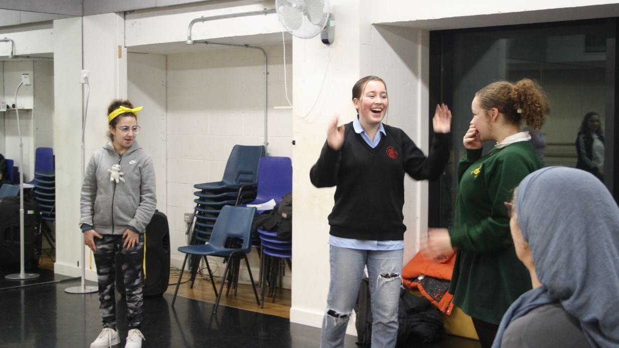 Three girls stand in a drama rehearsal space. Two of them stand looking at each with excited expressions.