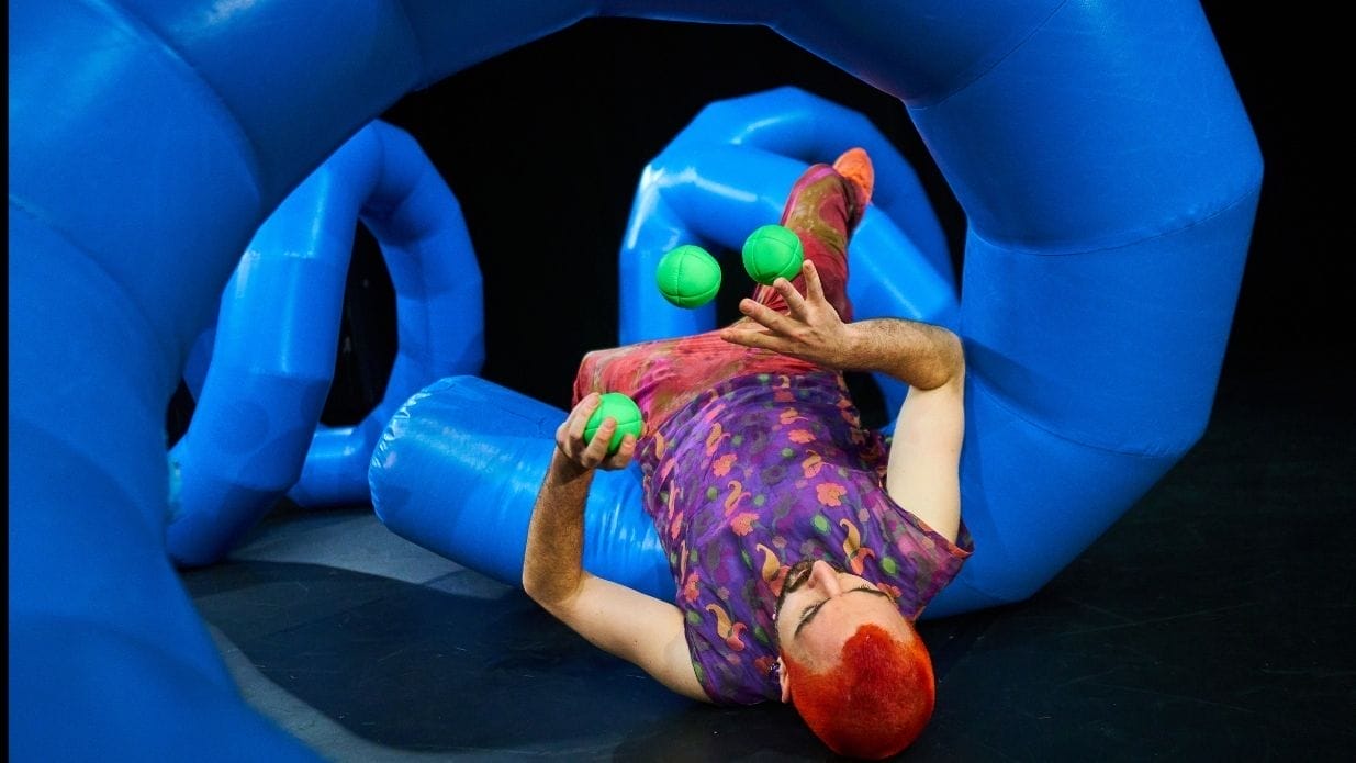 Juggler Wes Peden on stage. He is lying on his back surrounded by an inflatable blue structure and juggling three green balls.