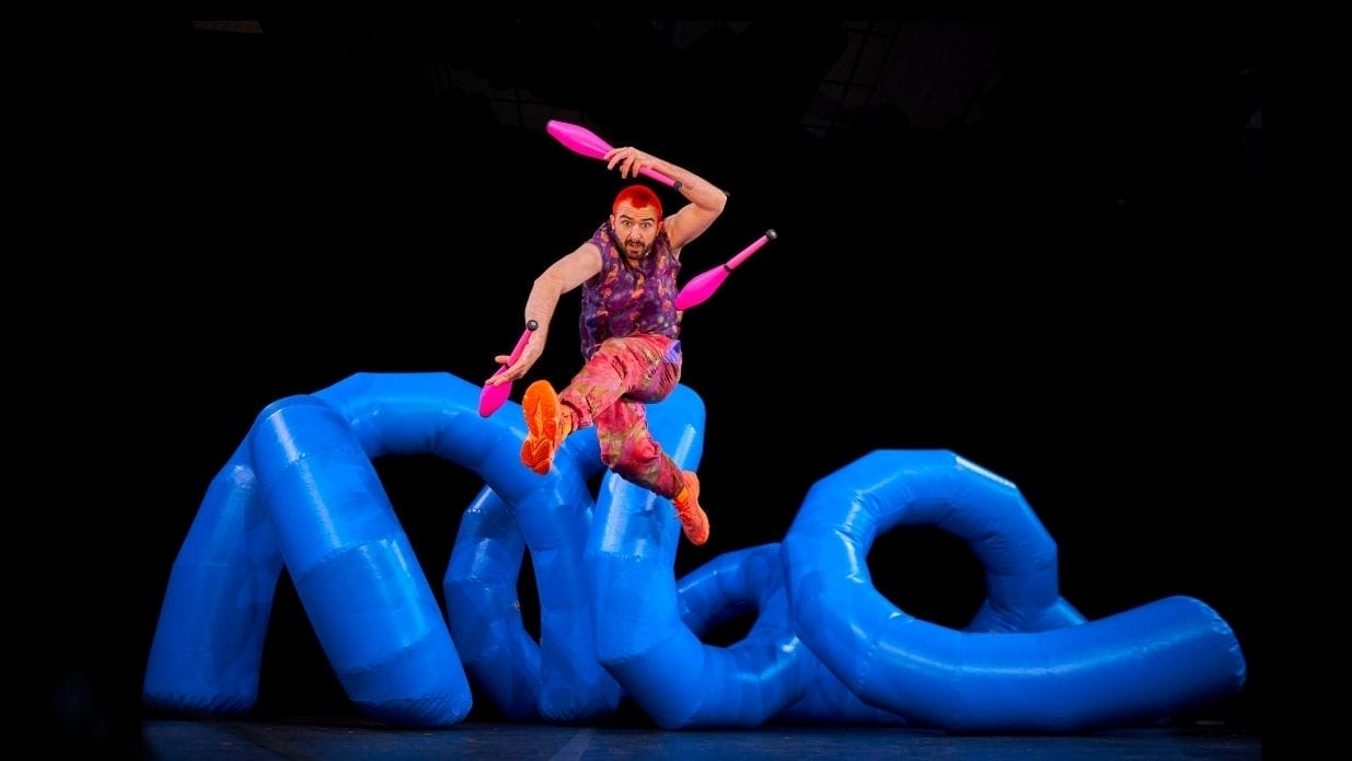 Juggler Wes Peden on stage. He leaps in the air juggling with three pink clubs. Behind him is an inflatable blue structure.
