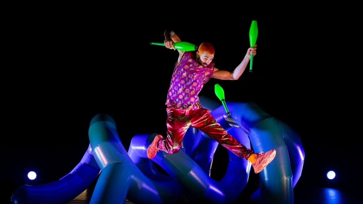 Juggler Wes Peden on stage. He leaps in the air juggling with three green clubs. Behind him is an inflatable blue structure.