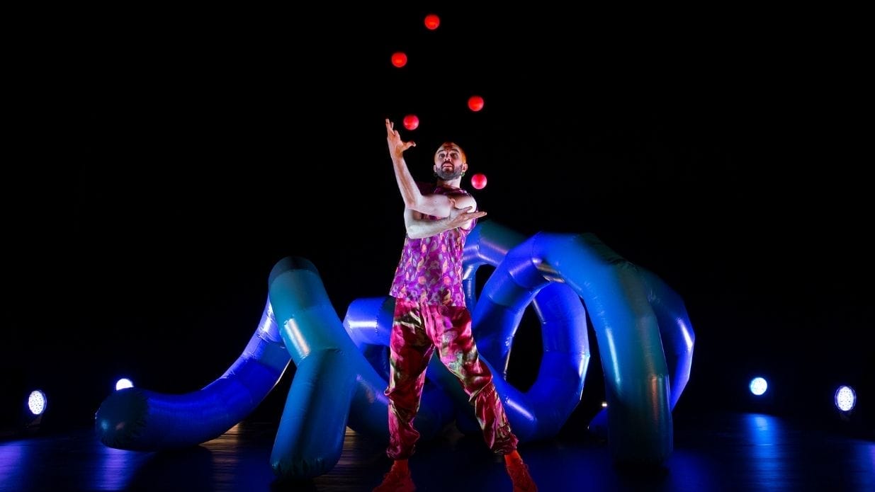 Juggler Wes Peden on stage. He is juggling with five pink balls in front of an inflatable blue structure.