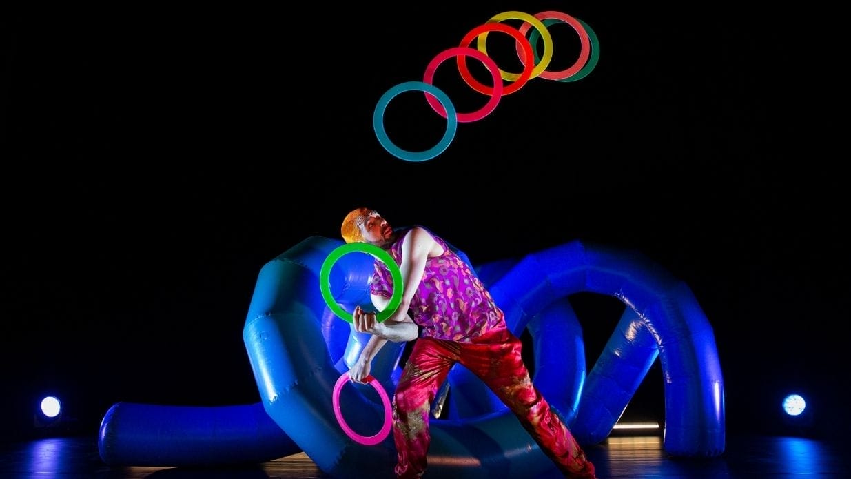 Juggler Wes Peden on stage. He juggles with 8 brightly coloured rings in front of an inflatable blue structure.