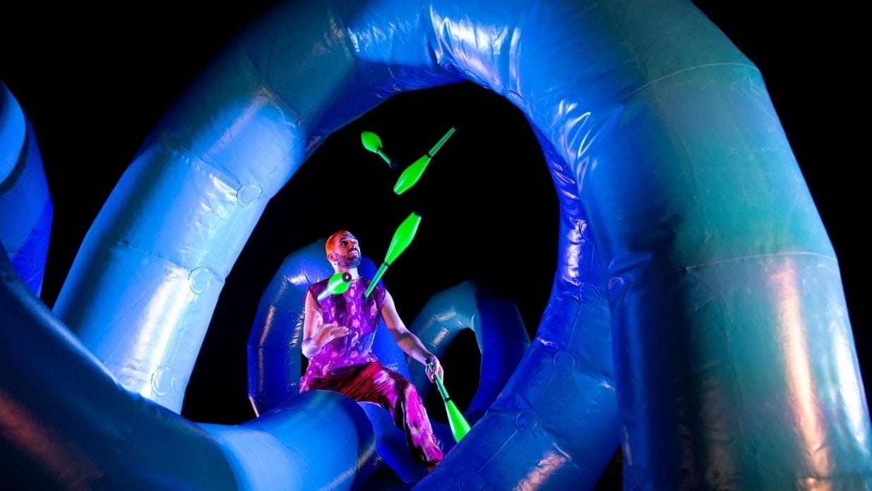 Juggler Wes Peden on stage. He is sitting on an inflatable blue structure and juggling 5 bright green clubs.