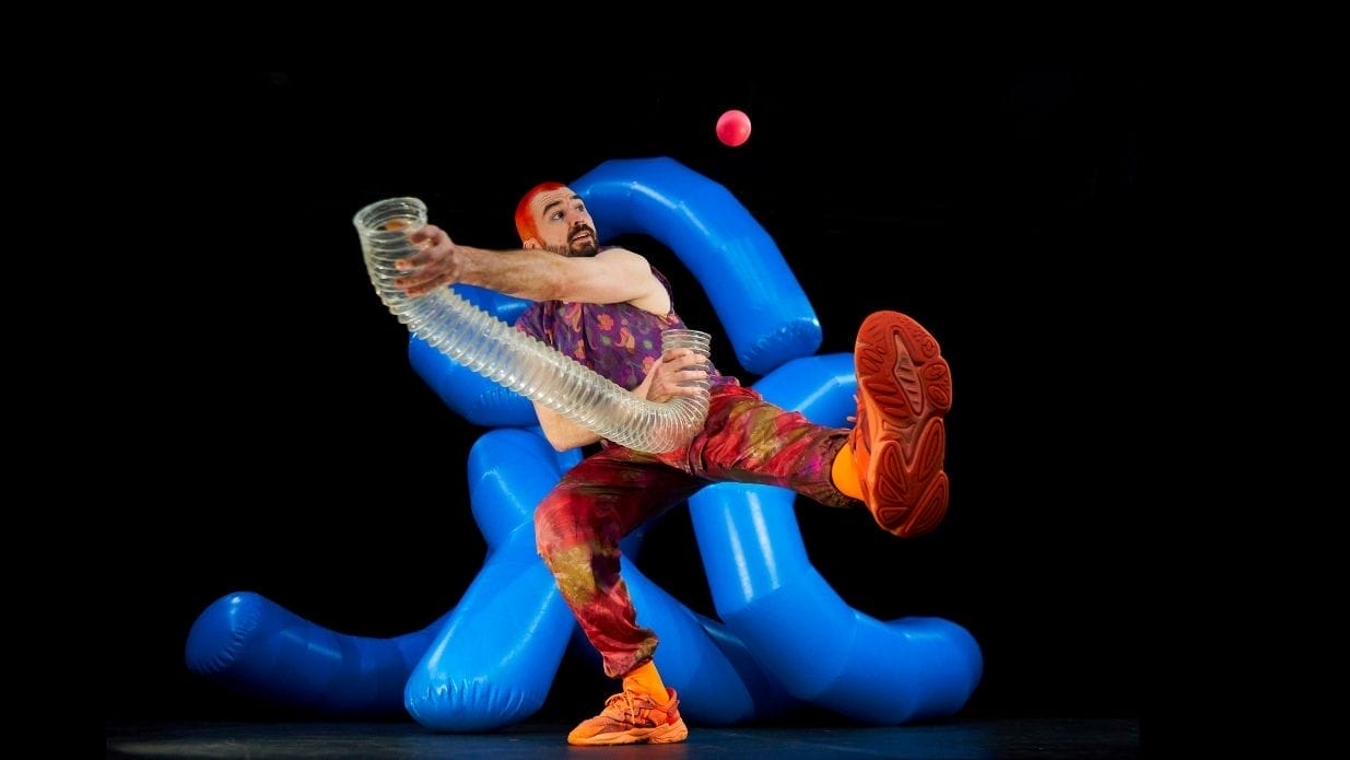 Juggler Wes Peden on stage. He stands on one leg holding a translucent tube which a pink ball has flown out of. Behind him is an inflatable blue structure.