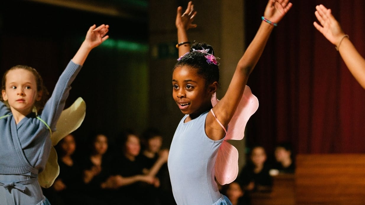 Two young girls taking part in a ballet workshop. They are wearing fairy wings with their arms in the air.