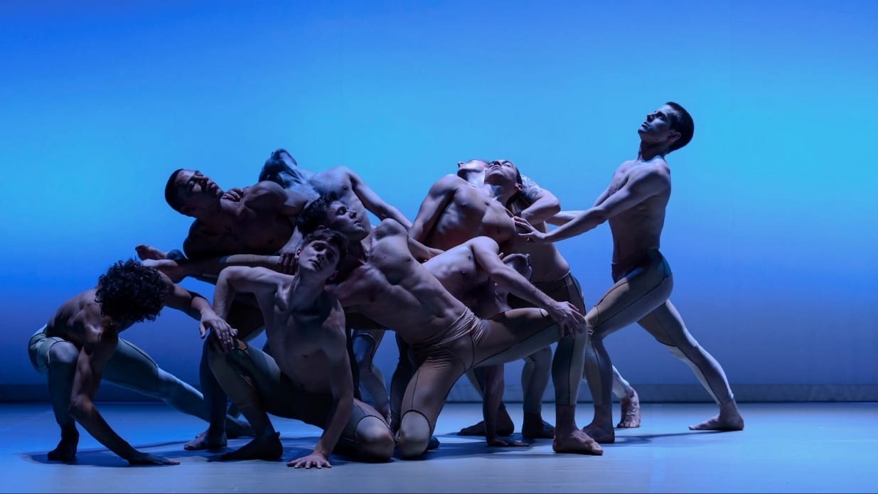 BalletBoyz on stage - a group of male dancers wearing pale coloured shorts are in a group, some kneeling or crouching, some standing. Connected by touch, they stretch in all directions.