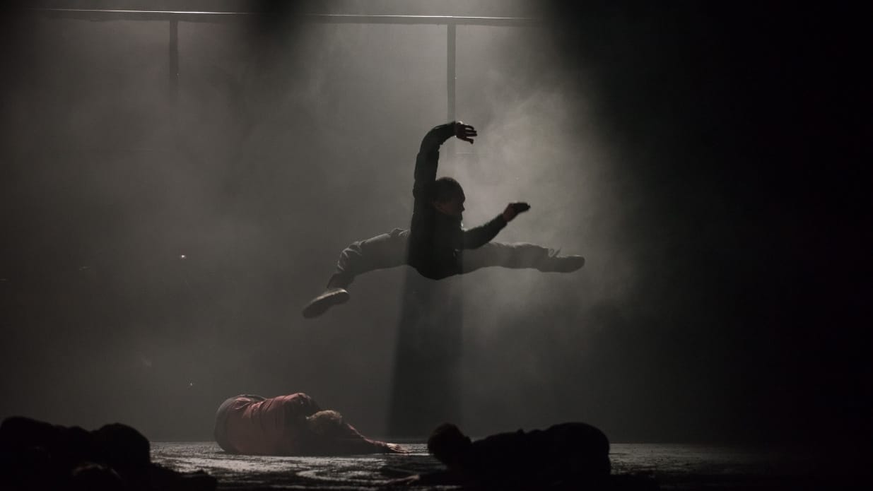 On a dark smokey stage, a dancer leaps with his back towards the floor. Around him, dancers lie on stage.