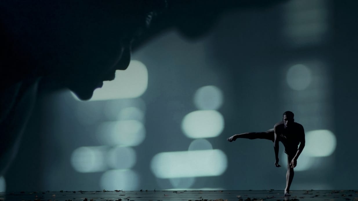 A dancer stands on one leg on a dark stage with lights in shades of green. In the foreground, a close up of a face in profile.