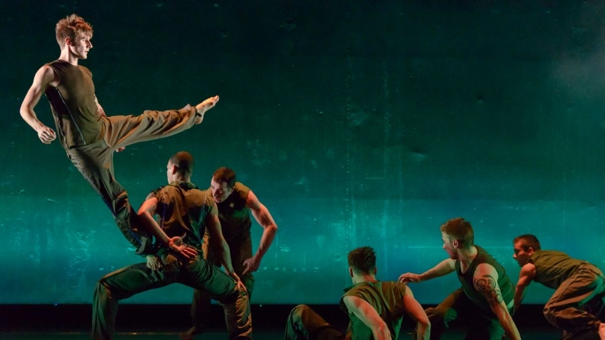 Dancers on stage in dark green costumes in front of a green backdrop. One stands on another's bent leg, about to fall sideways.