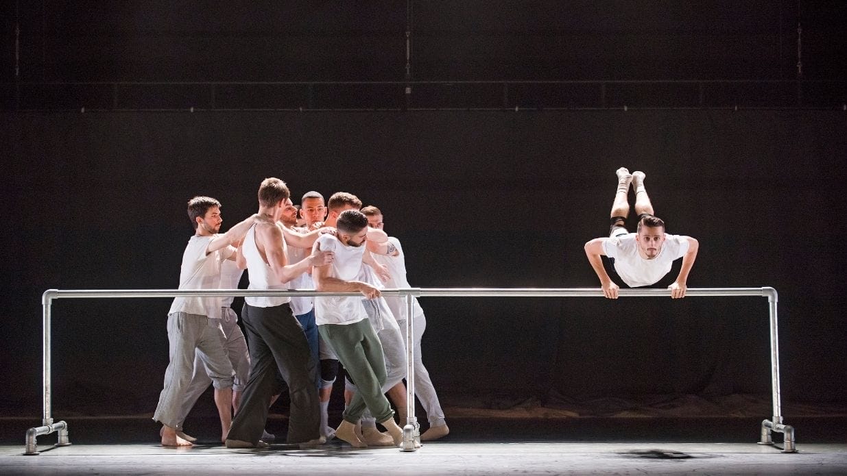 A group of dancers in white tshirts and grey trousers huddle together, jostling for space, while another leaps with his hands on a ballet bar.