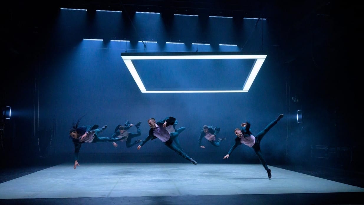 Five male dancers wearing suits leap sideways underneath a square of light