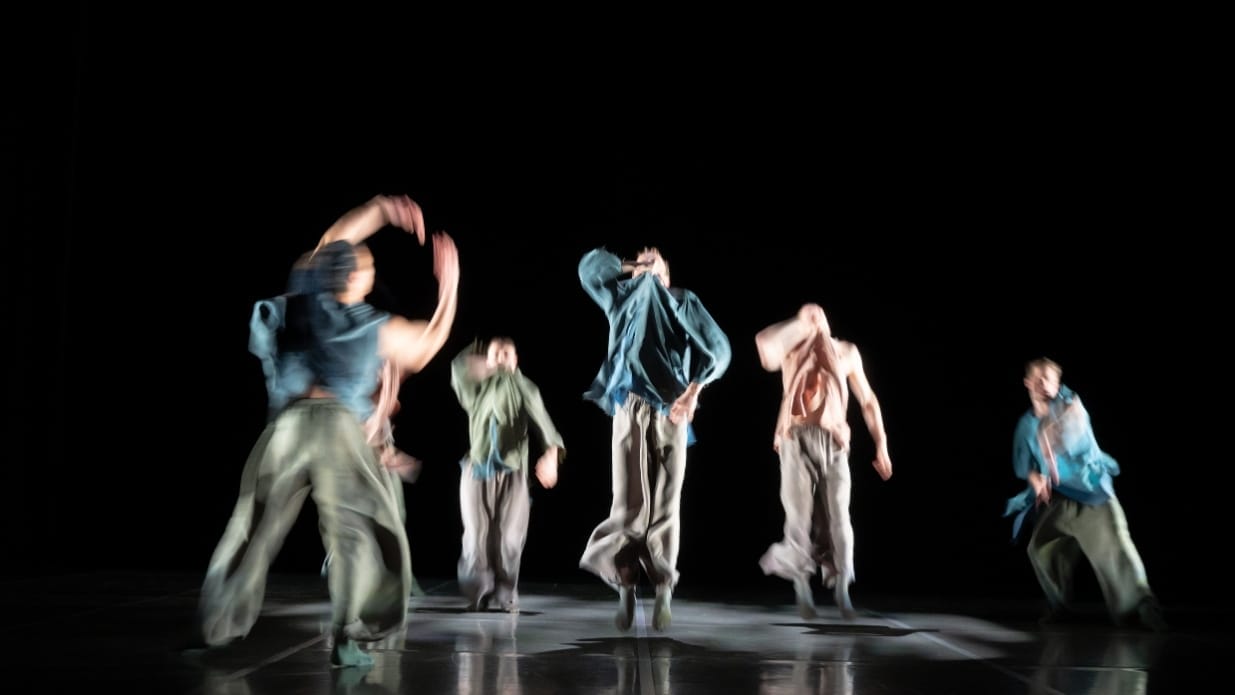 Five dancers on stage in Ripple, they wear loose clothing and their images are blurred
