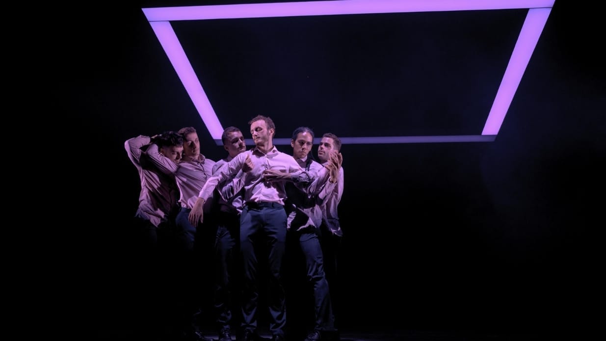 A group of 6 male dancers stand together, connected by touch. They are underneath a glowing light in the shape of a purple square.