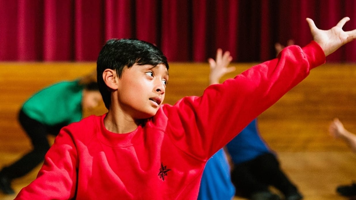 A group of young people taking part in a dance workshop. In the foreground a boy wearing a red sweatshirt stretches his arm to the side.