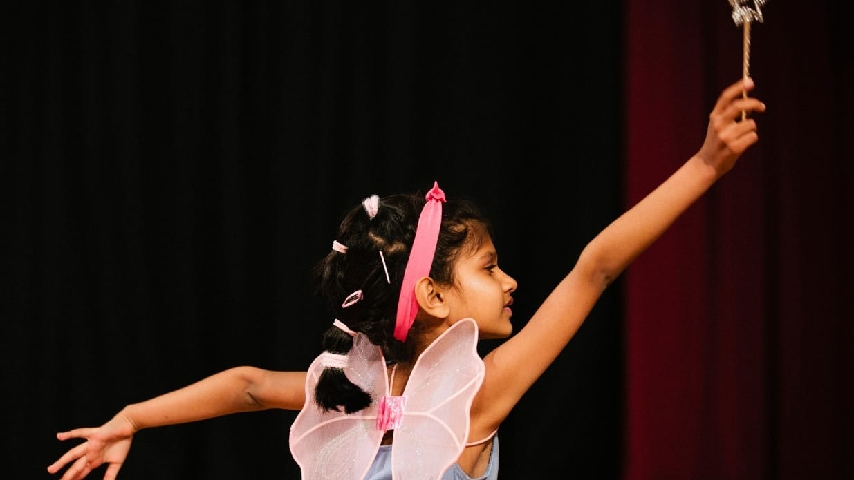 A girl wearing fairy wings and a pink headband holds a wand with a star on top.
